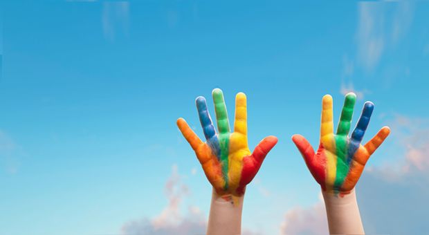 Childs hands covered with multicoloured paint reaching up to the blue sky