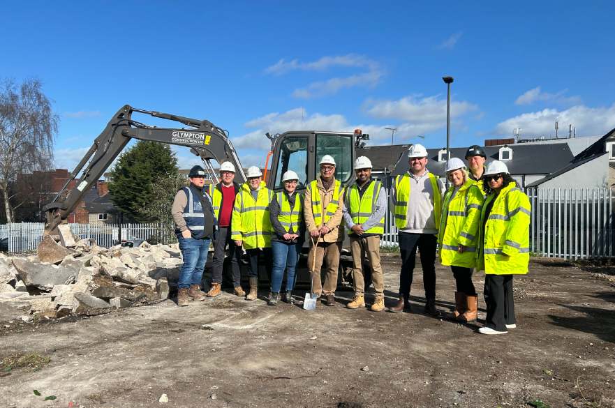 Contractors and Council officers gather around a spade to mark the start of work on affordable homes to rent, with a digger in the background.