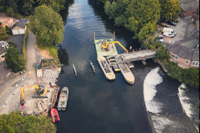 A large excavator on a floating pontoon on a river, next to a half demolished bridge.