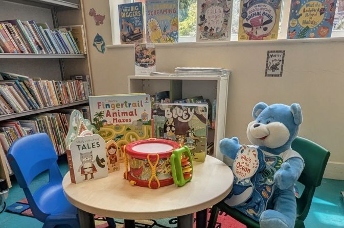 The children's area of a library featuring bookshelves and a small table and chairs with books and toys