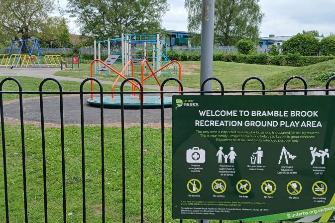 A view of a playground seen through the perimeter fence showing new play equipment. There is a sign on the fence that reads Bramble Brook Recreation Ground
