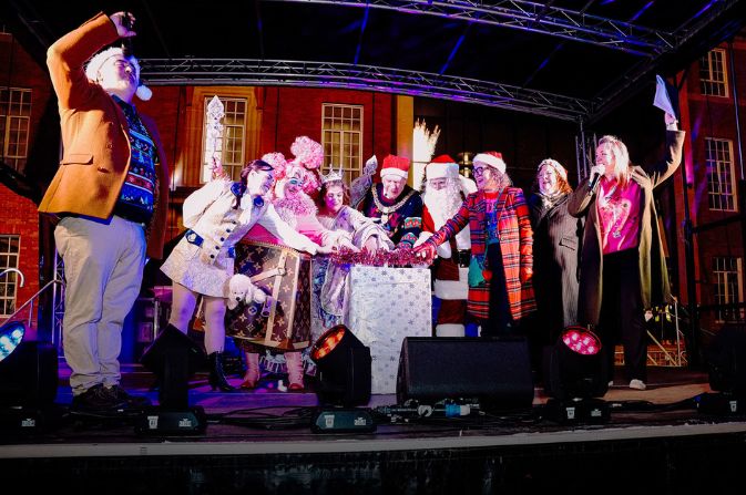 A group of people including panto stars and councillors standing on a stage push the button to turn on Derby's Christmas lights