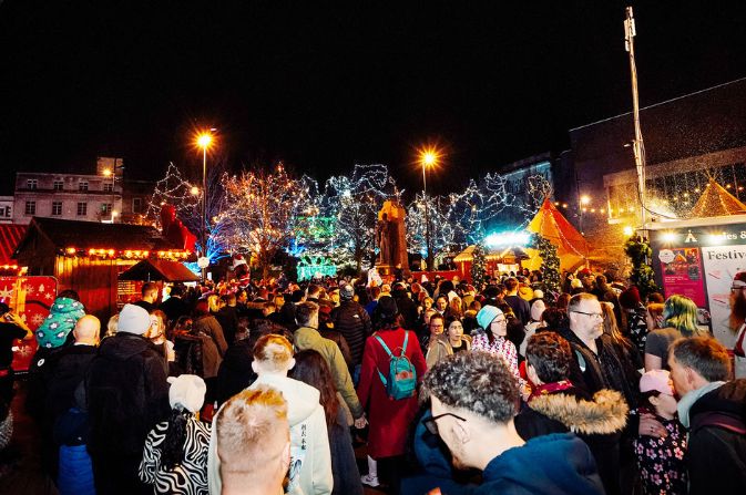 A crowd of people gather in Derby Market Place at night against a backdrop of trees decorated with Christmas lights
