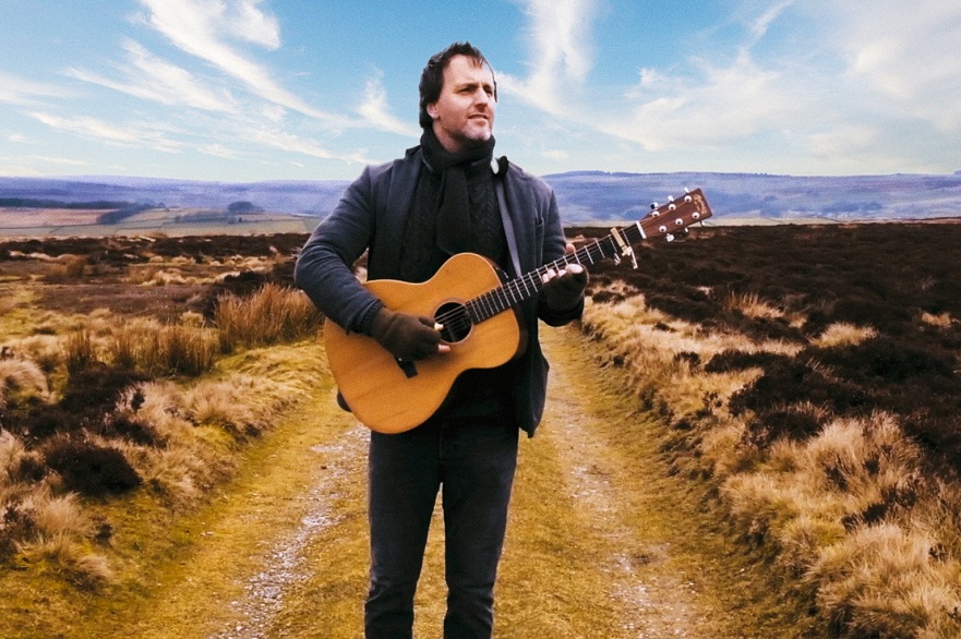 Former Bellowhead frontman Jon Boden standing alone on a dirt road with a wide blue sky in the background