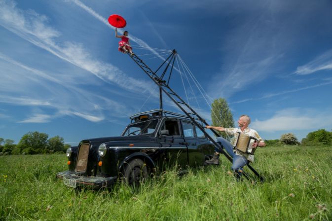 A performer holding a red umbrella sits high up at the top of a metal structure which is secured to a black taxi cab. Another performer sits on the bottom of the structure.