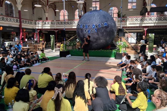 A man speaking on a stage inside Derby Market Hall surrounded by seated children watching him