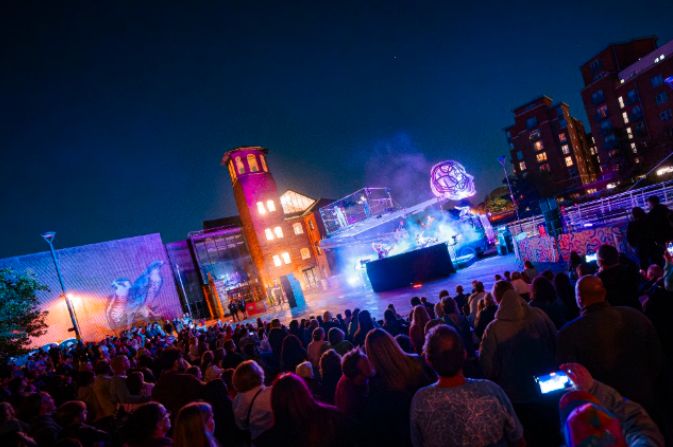 Crowds watch a night time performance taking place on a huge illuminated aerial platform outside the Museum of Making in Derby
