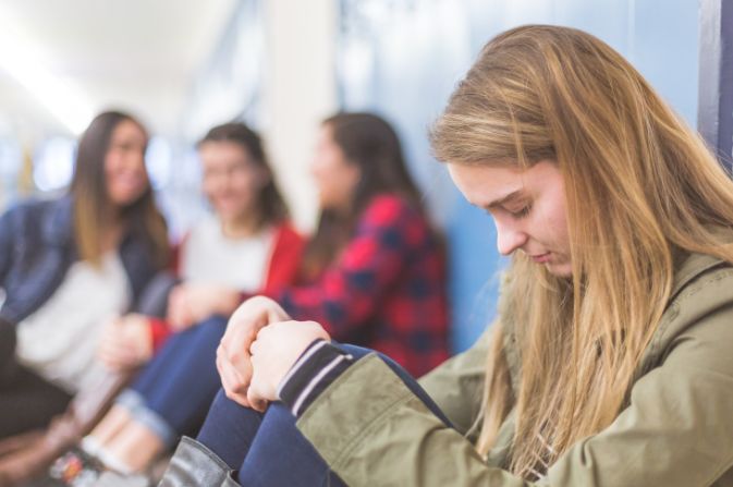 A teenage girl looks down as three girls in the background talk about her