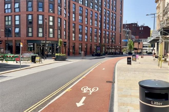 A red cycle lane adjacent to a recently paved road. The road is running alongside a large apartment building.