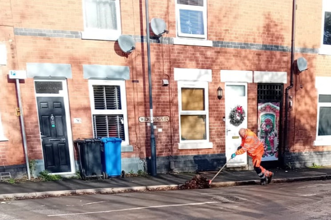 The image shows a council worker sweeping leaves from the kerb.