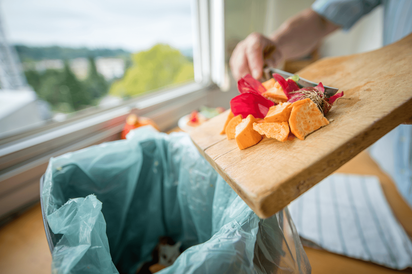 The image shows someone scraping food waste from a wooden chopping board into a small caddy on the worktop.